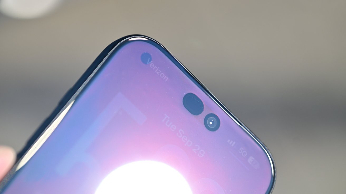 Close-up of a smartphone's top screen showing pill-shaped front camera cutout, glossy edges, and reflection of bright light on the display
