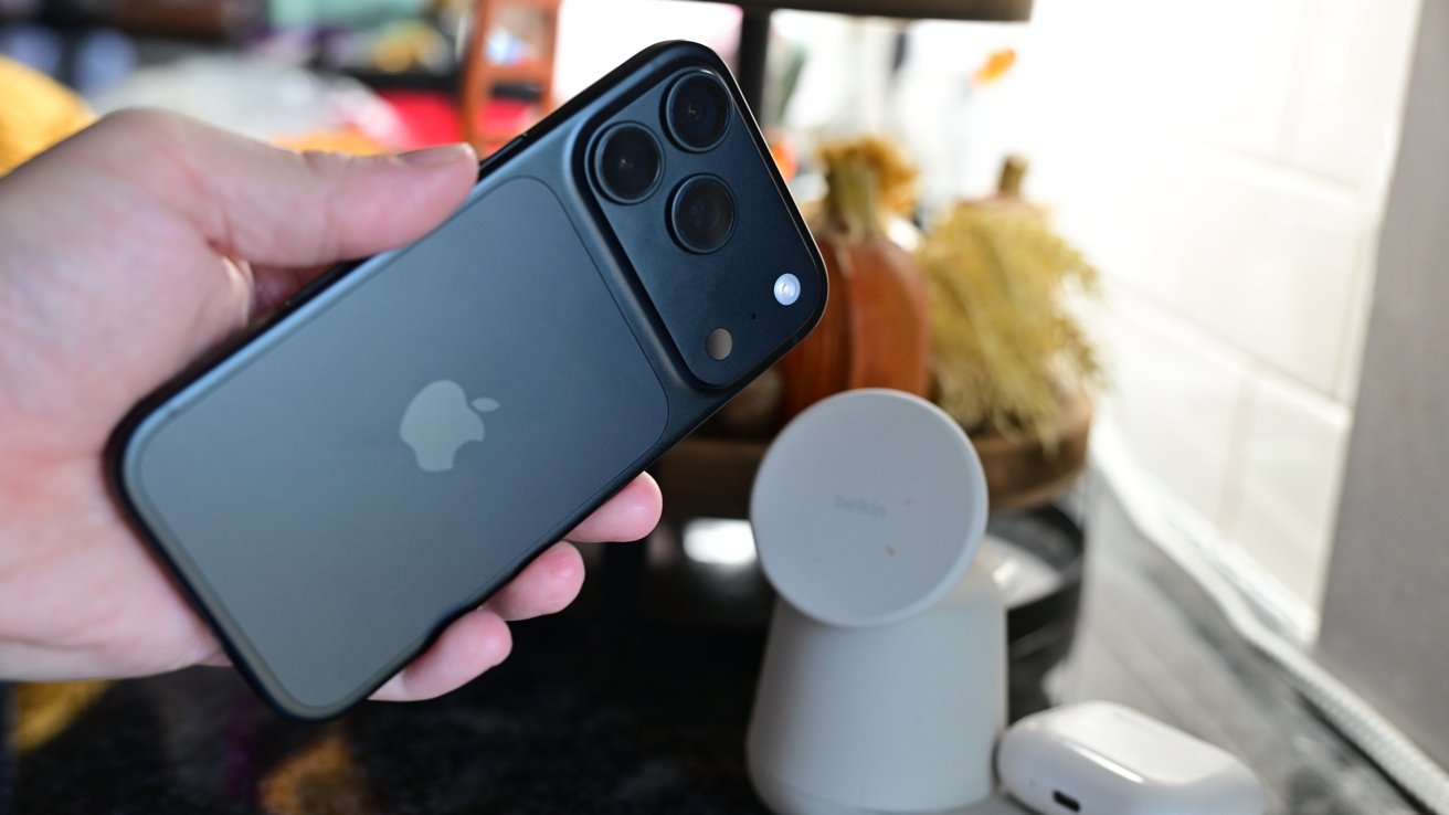 Hand holding a dark-colored iPhone with triple camera system, near a white wireless charger and AirPods case on a kitchen counter with blurred autumn decorations in the background