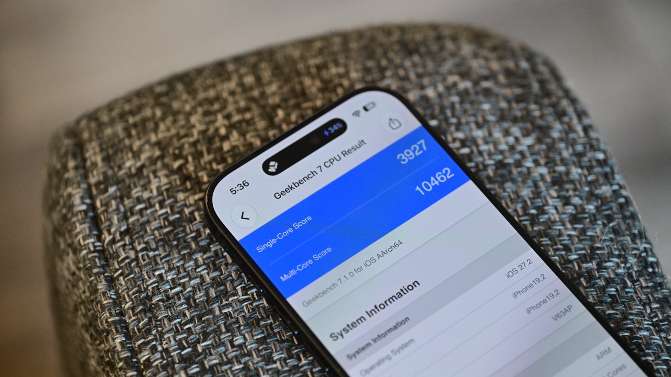 Smartphone resting on a textured gray cushion, screen displaying a blue and white benchmarking or system information app with performance scores and device details visible