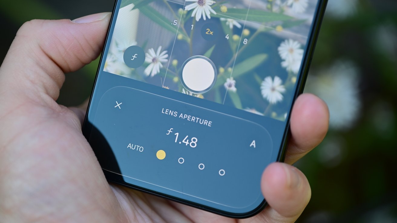 Hand holding a smartphone camera focused on white flowers, with on-screen controls showing manual lens aperture adjustment set to f1.48 and various camera settings visible at the bottom