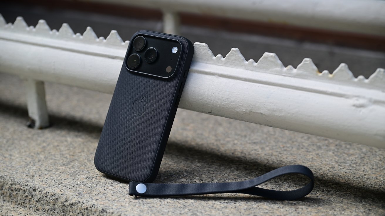 Black smartphone with triple rear cameras in a dark case, propped against a white metal railing on concrete, with a matching black wrist strap lying beside it