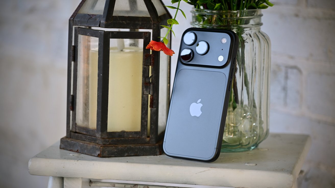 Modern iPhone with dual rear cameras leaning against a rustic lantern holding a candle, beside a glass jar of green plants, all arranged on a small white wooden table