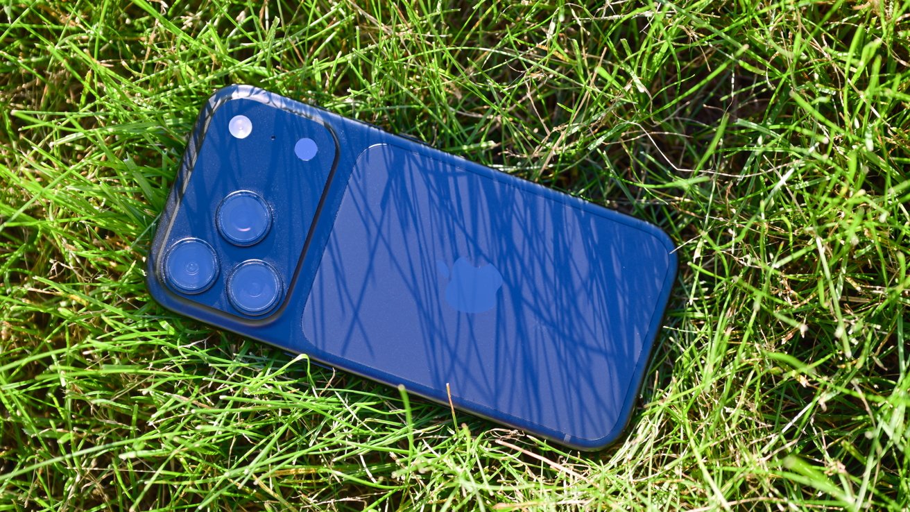 Blue smartphone lying face down on bright green grass, showing triple camera lenses and glossy back reflecting thin blades of grass
