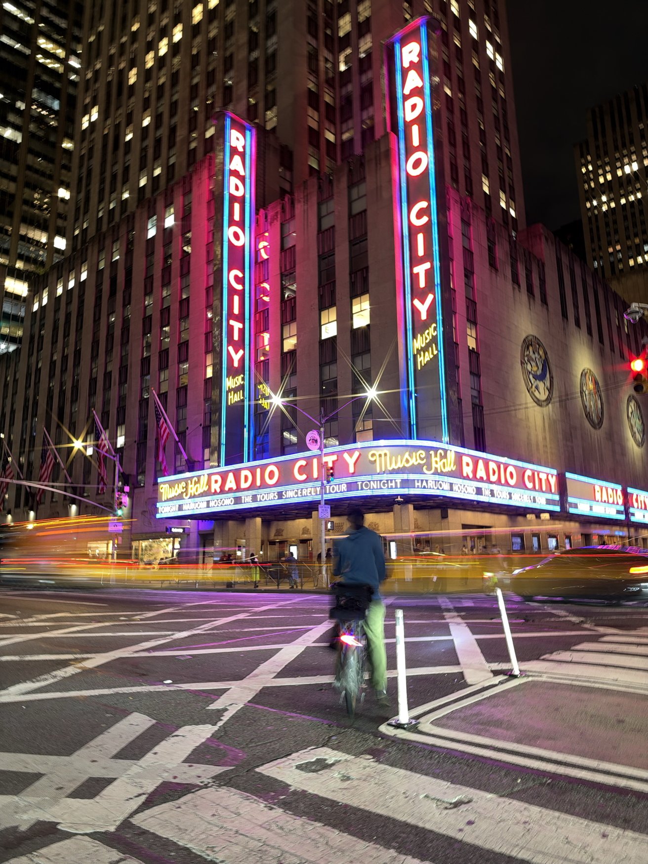 Nighttime street scene at Radio City Music Hall, neon signs glowing pink and blue, tall surrounding buildings, blurred car lights, and a cyclist crossing the wide striped intersection