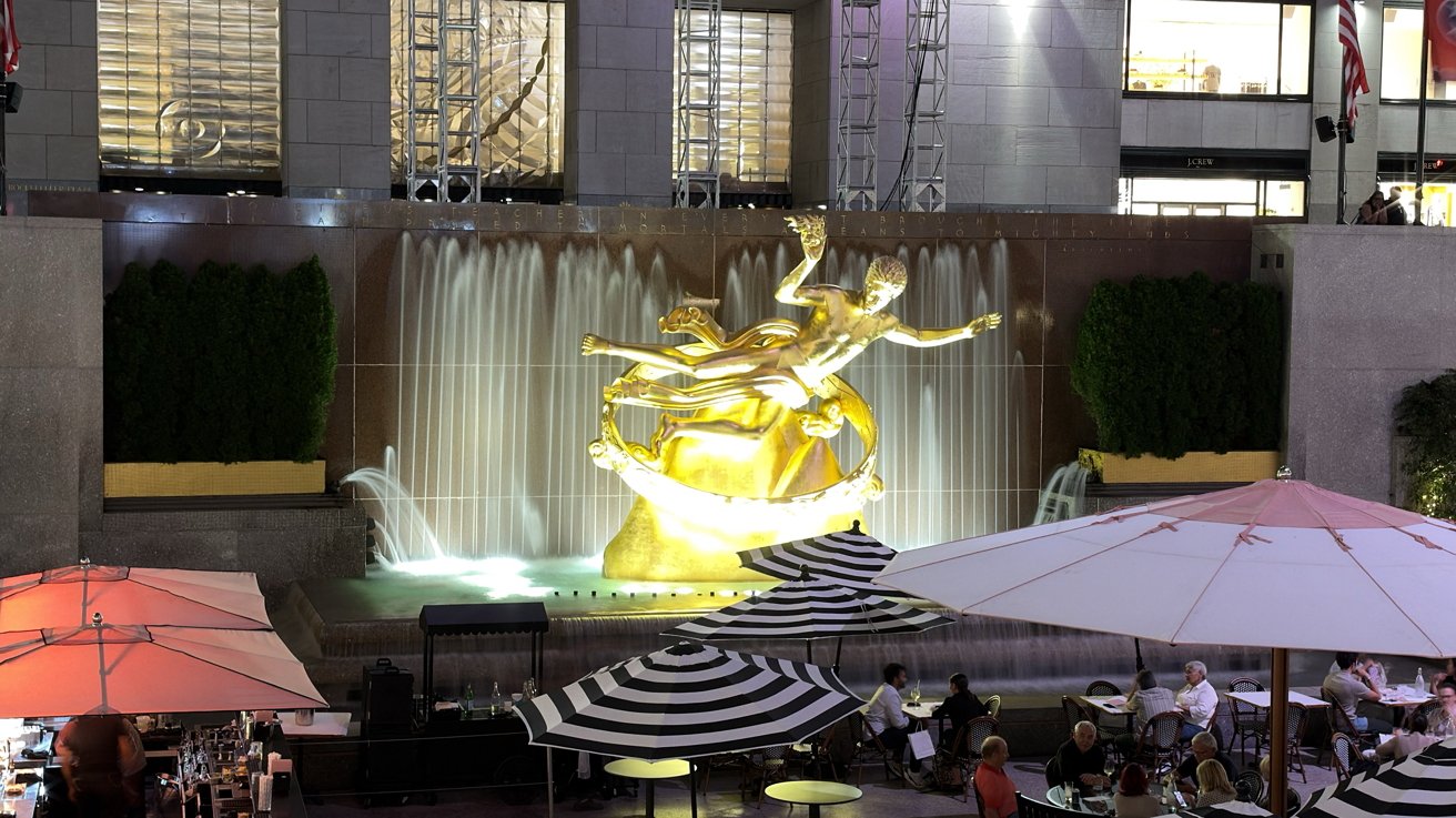 Golden Prometheus statue before cascading fountain at Rockefeller Center, surrounded by outdoor dining tables, striped umbrellas, and evening lights with people seated and walking nearby