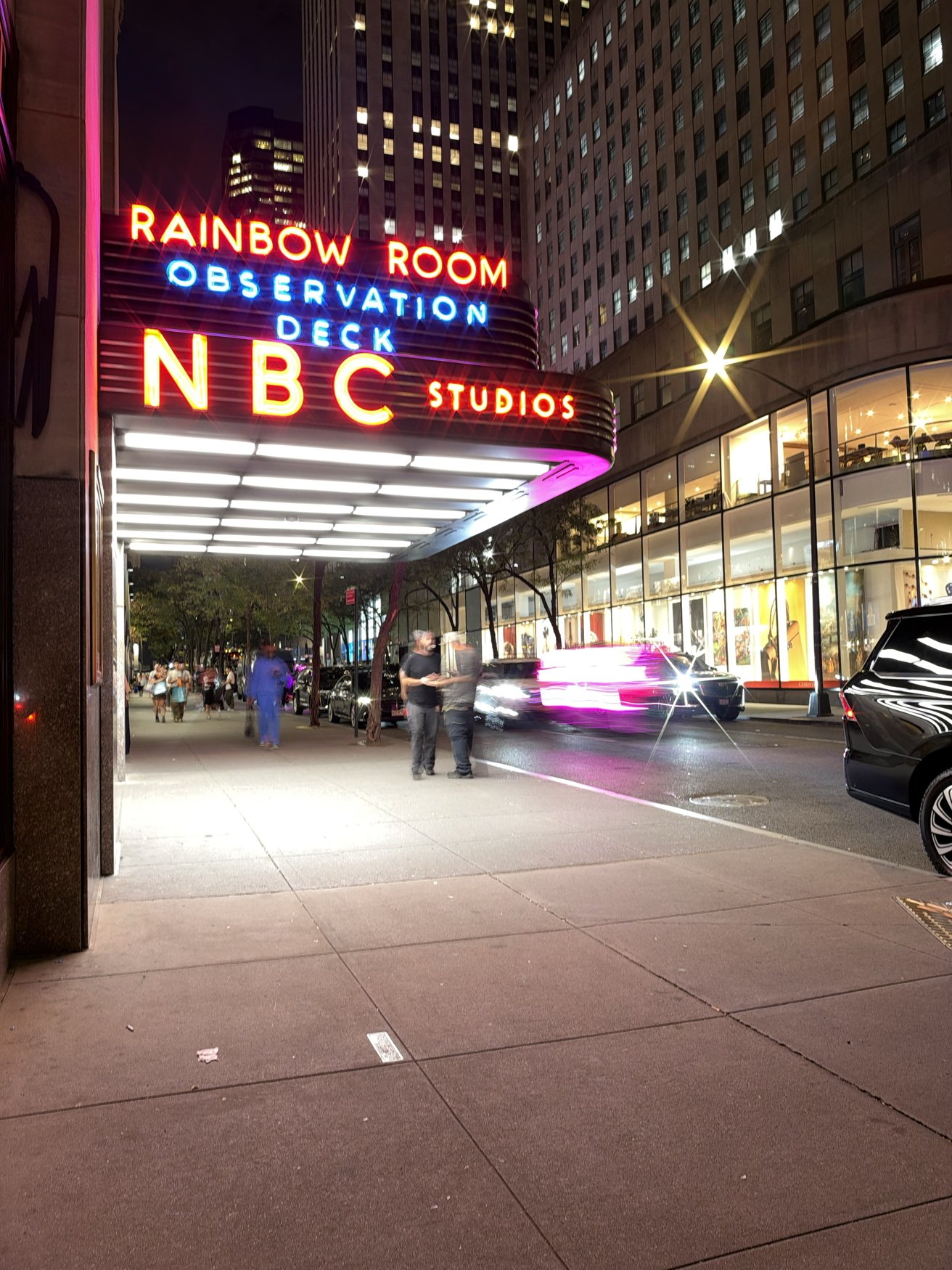 Night street scene at NBC Studios entrance, bright neon Rainbow Room Observation Deck sign overhead, pedestrians walking under canopy, blurred car lights and tall office buildings surrounding the sidewalk