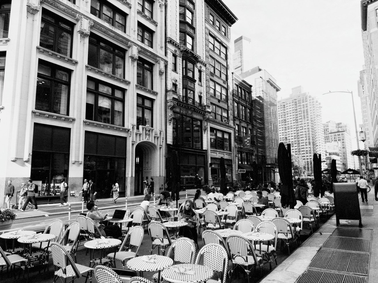 Outdoor cafe tables and chairs line a busy city sidewalk beside tall historic buildings, with people walking and dining in a lively urban street scene, shown in black and white