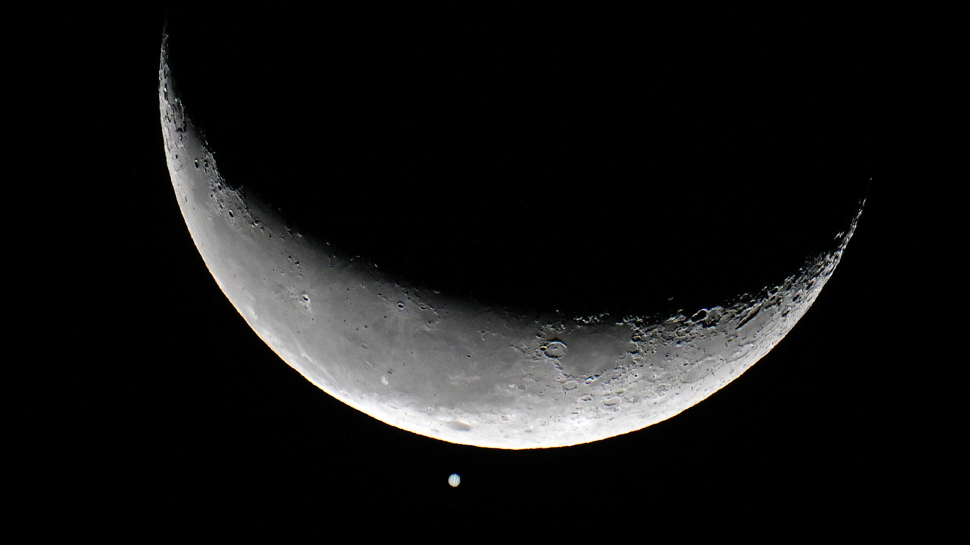 The crescent moon is shown in a black night sky with a bright point of light representing Jupiter positioned below its sunlit crescent.