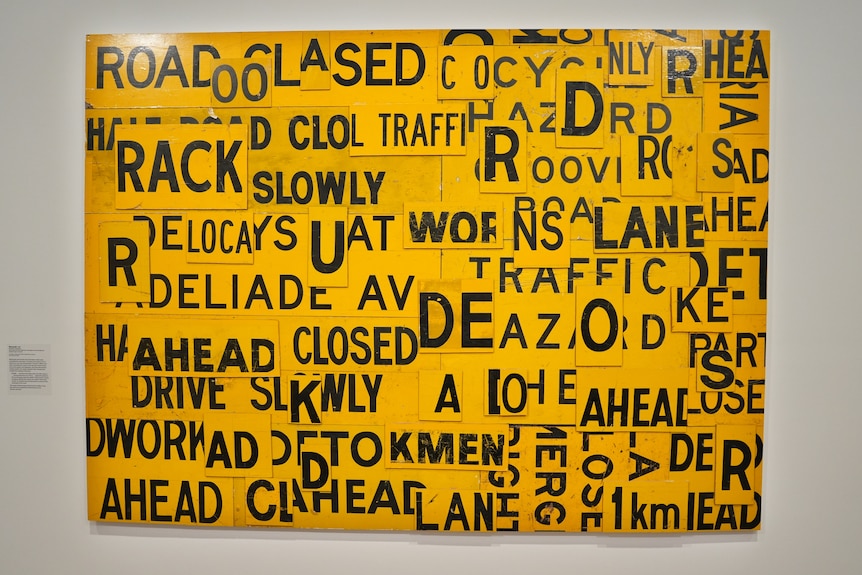 Artwork that is yellow and includes road signs mixed together including words like 'ahead' and 'slowly'.