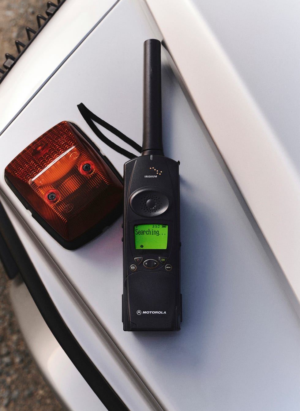 Satellite phone displaying "Searching..." next to a red emergency light on a vehicle's surface. Satellite phone displaying "Searching..." next to a red emergency light on a vehicle's surface.