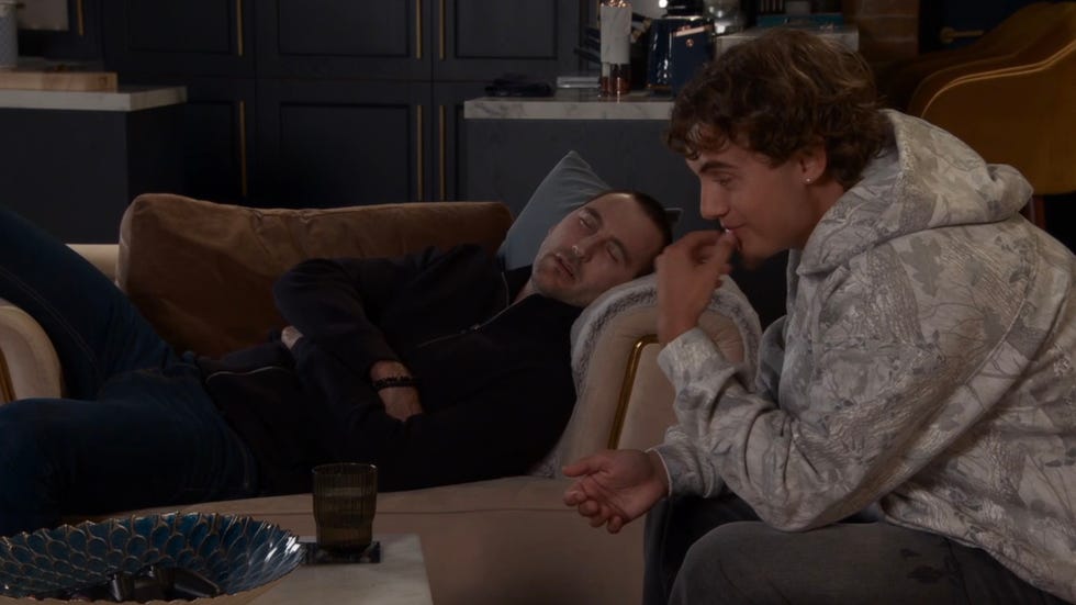 person sleeping on a couch while another person sits nearby, smiling slightly. person sleeping on a couch while another person sits nearby, smiling slightly.