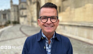 Justin Hunter smiles at the camera, he wears a blue labourers jacket with a blue floral shirt underneath and black round glasses. He is standing outside Gloucester Cathedral