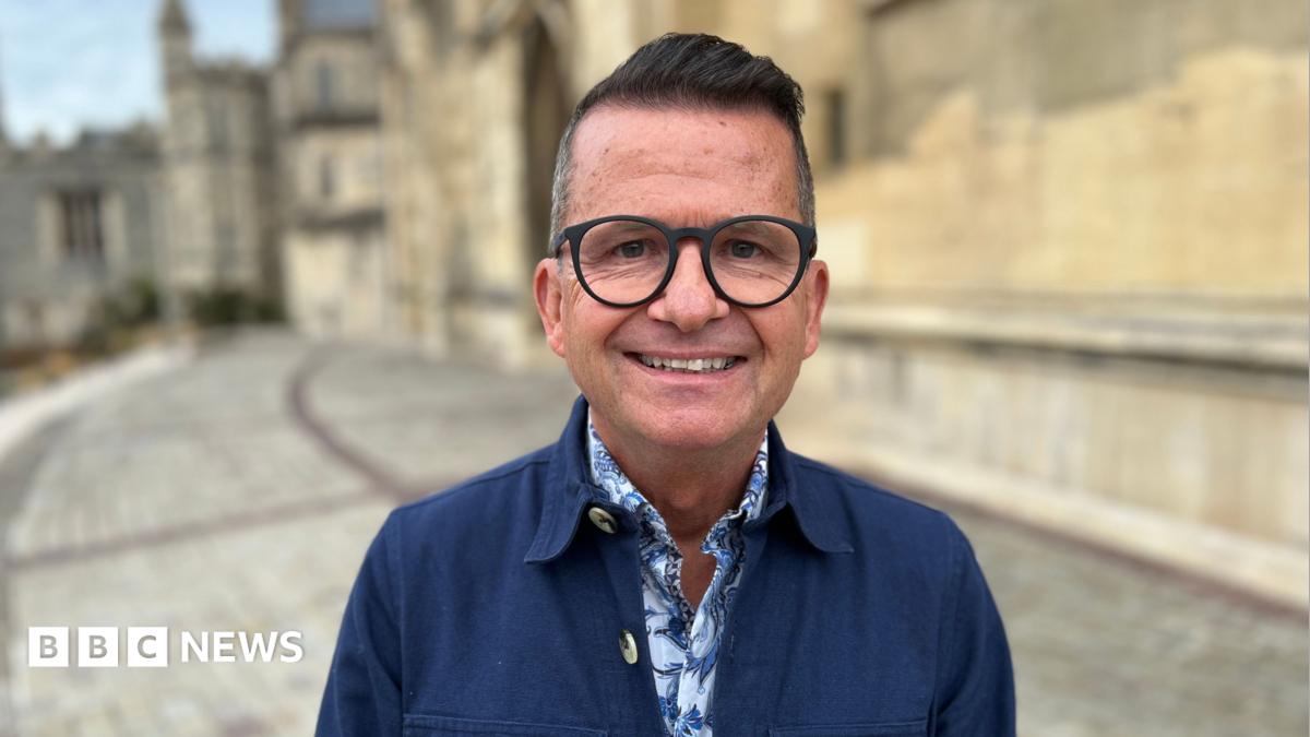 Justin Hunter smiles at the camera, he wears a blue labourers jacket with a blue floral shirt underneath and black round glasses. He is standing outside Gloucester Cathedral