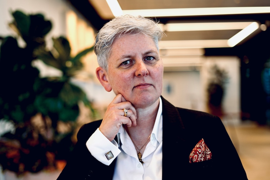A woman with short grey hair, wearing a dark suit jacket over a white shirt, looks intently into the camera.