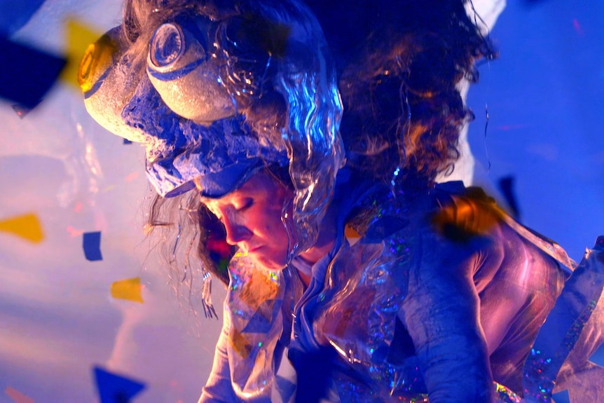 A still of a woman in an ostentatious costume, looking upset. She looks down, and her headpiece includes two upturned bowls.