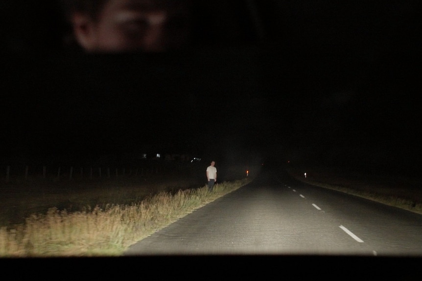 a wide shot of a man in white stood on a road at night