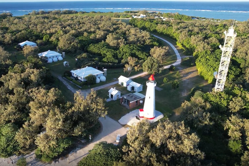 An aerial view of a lush island, featuring a lighthouse, three houses and a transmission tower.