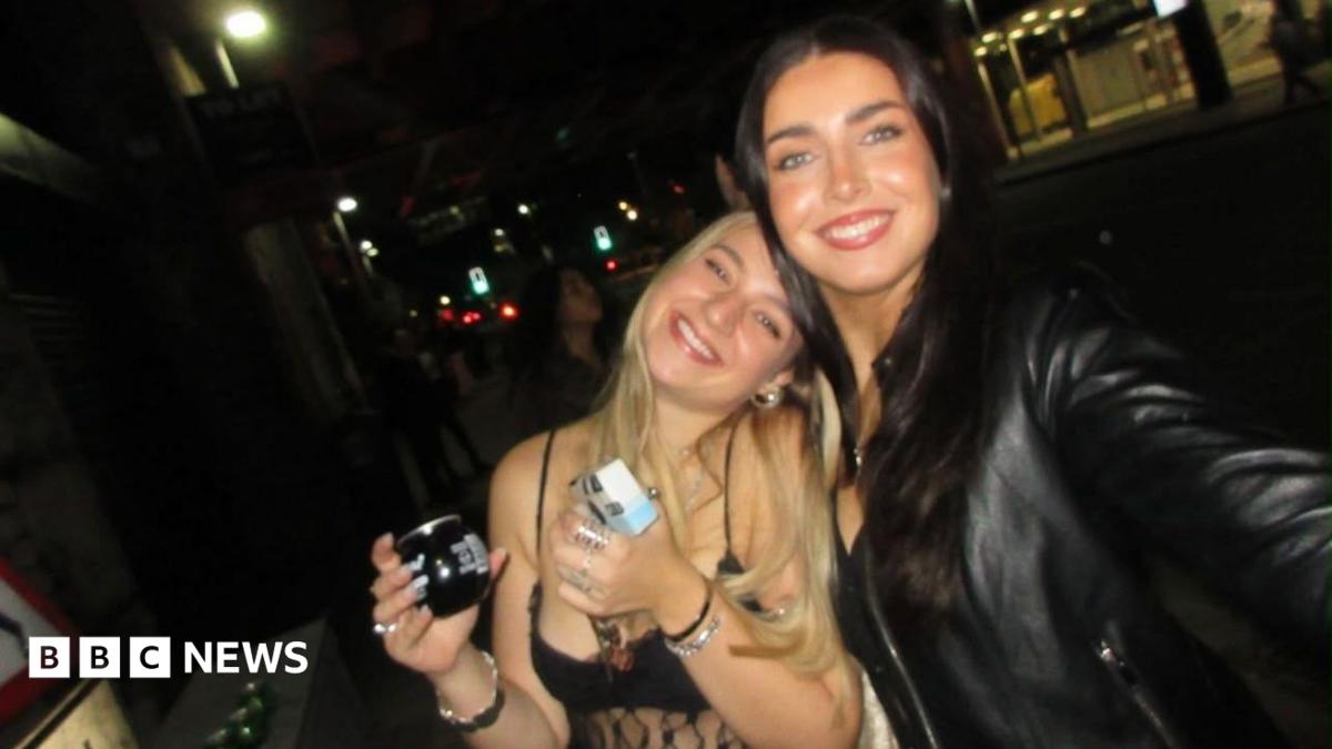 There are two girls in a dark bar smiling at the camera . On the left a blonde girl is wearing a strappy top and holding a drink. She leans into her friend on the left who is holding the camera to take a selfie of the two of them. The girl on the right has long brown hair and is wearing a black leather jacket.