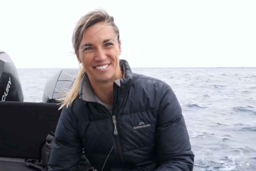 A woman with blonde hair sitting on a boat smiling, with the ocean in the background