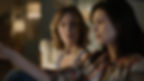 Two women sit indoors; one in focus looks concerned at the other in a plaid shirt in the foreground, with a warm, dim background.