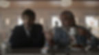 Two men sit at a diner counter; one gestures with a mug while talking, the other listens. Dim, smoky interior with a tense mood.