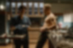 Nurse in scrubs examines a small card while a shirtless man stands beside her in a tense hospital prep room.