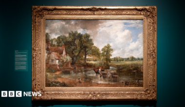 A large oil painting displayed on a wall as part of an art exhibition. The painting depicts a rural scene with a horse and cart moving through a river. There is a cottage on the bank of the river.