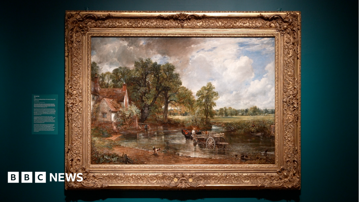 A large oil painting displayed on a wall as part of an art exhibition. The painting depicts a rural scene with a horse and cart moving through a river. There is a cottage on the bank of the river.