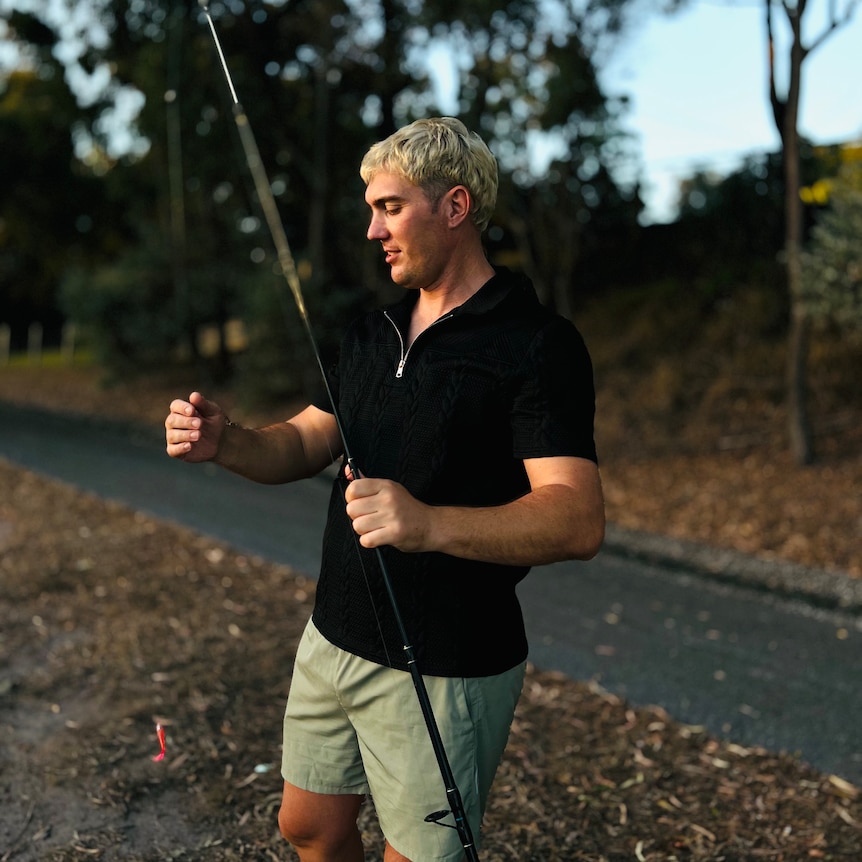 A man stands outdoors by a path. He is looking down at the fishing rod he is holding.