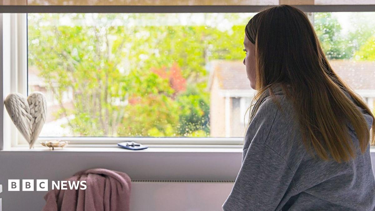 A young woman looks out of her bedroom window in a stock image. Outside there are trees visible and in the room the wall is a pale purple with a heart-shaped wing ornament on the windowsill.
