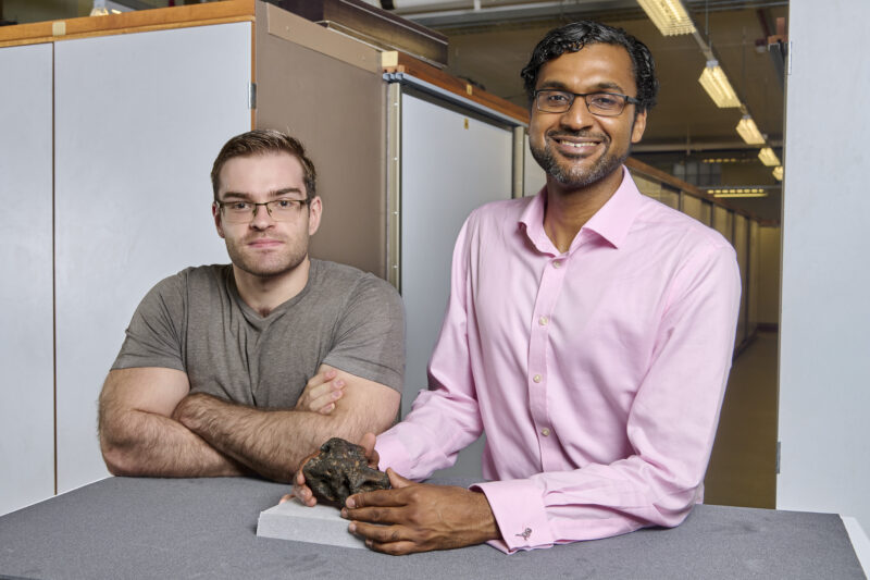 A man at left with arms folded and glasses, a man at right with pink button up shirt and glasses holding a fossil.