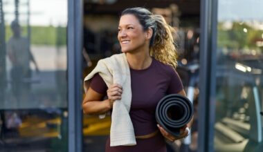 Woman setting up for boat pose, holding yoga mat and towel, coming out of studio smiling