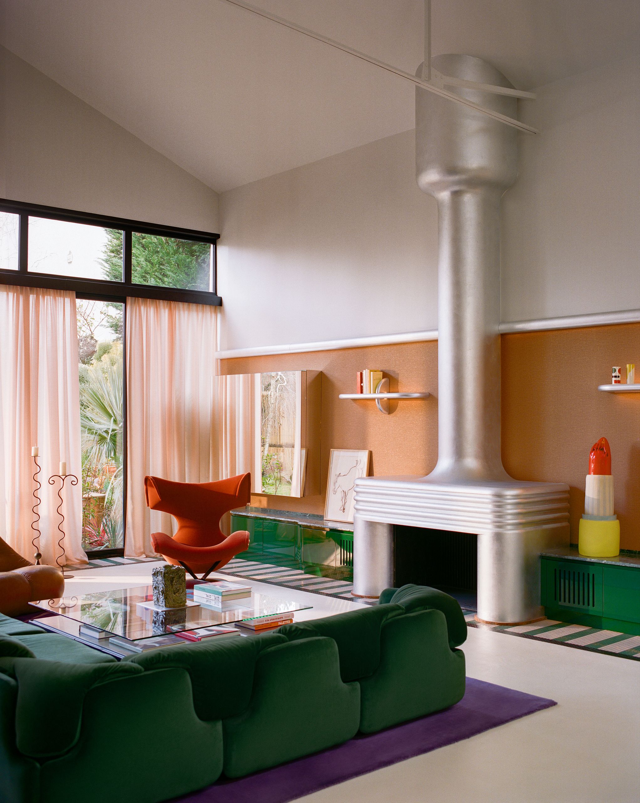 A colorful, Space Age-inspired living room with a dark green sofa, a purple rug, a red reading chair, orange wall paper, pink sheer curtains, and a brushed chrome fireplace.