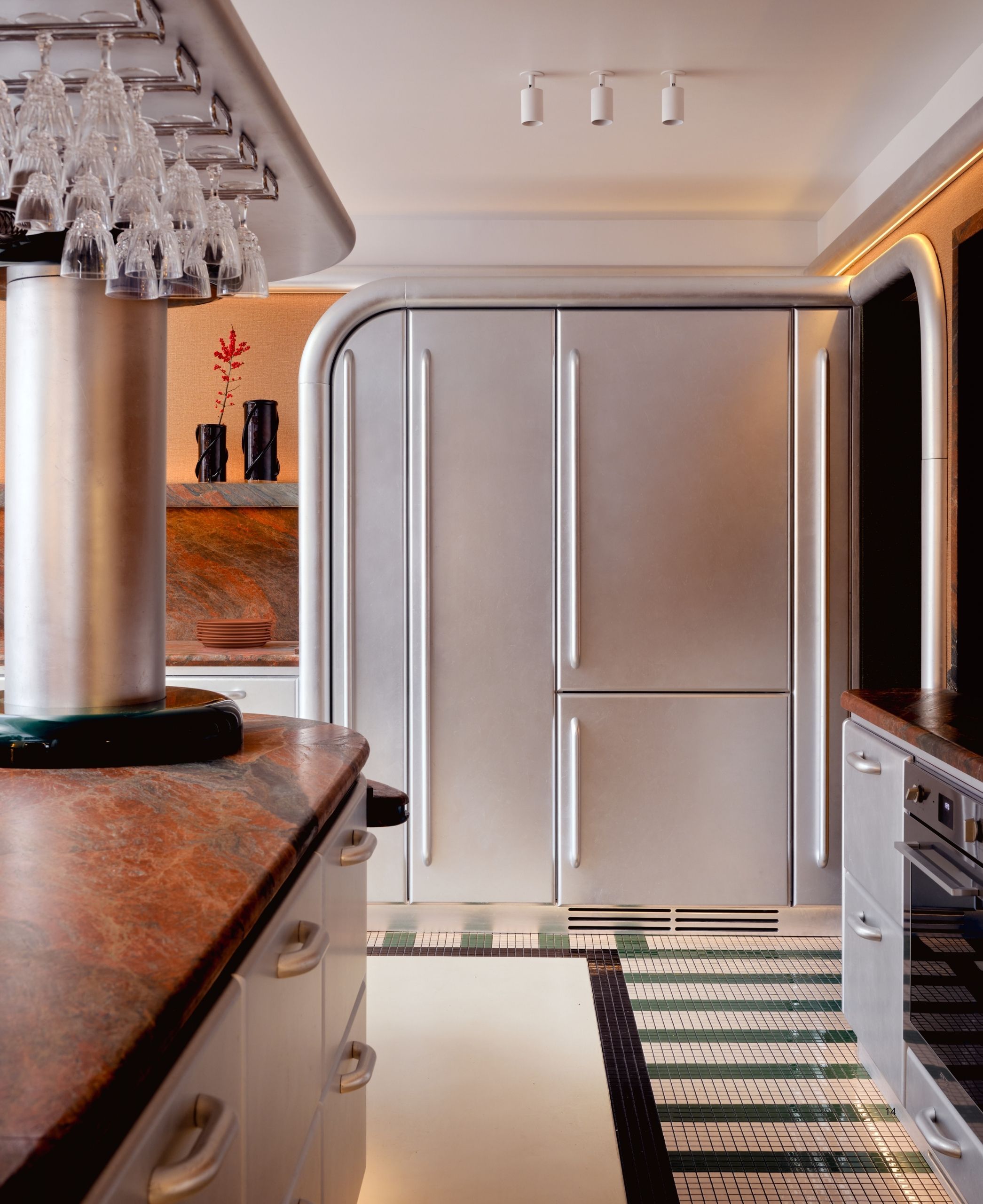A Space Age-inspired kitchen with a curved, brushed chrome, large fridge. The floors are green mosaic tiles, and there is an island with red marble and glasses hanging over it.