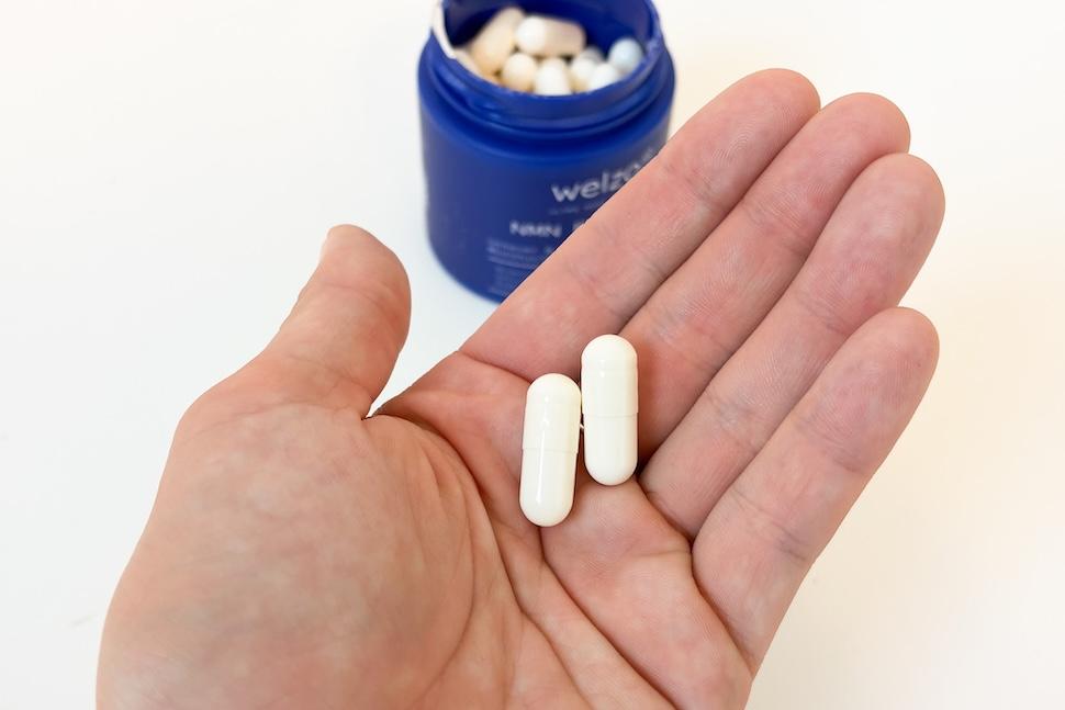 Closeup of an open palm holding two off-white capsules of Welzo Ultra Purity NMN Pro 1000. You can see an open canister of capsules on the countertop below.