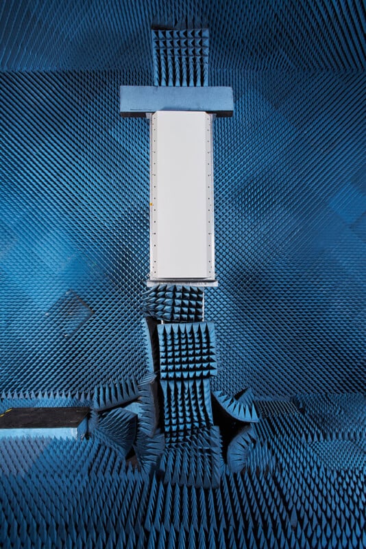 A rectangular white device is mounted vertically in the center of an anechoic chamber lined with blue, pyramid-shaped foam absorbers.