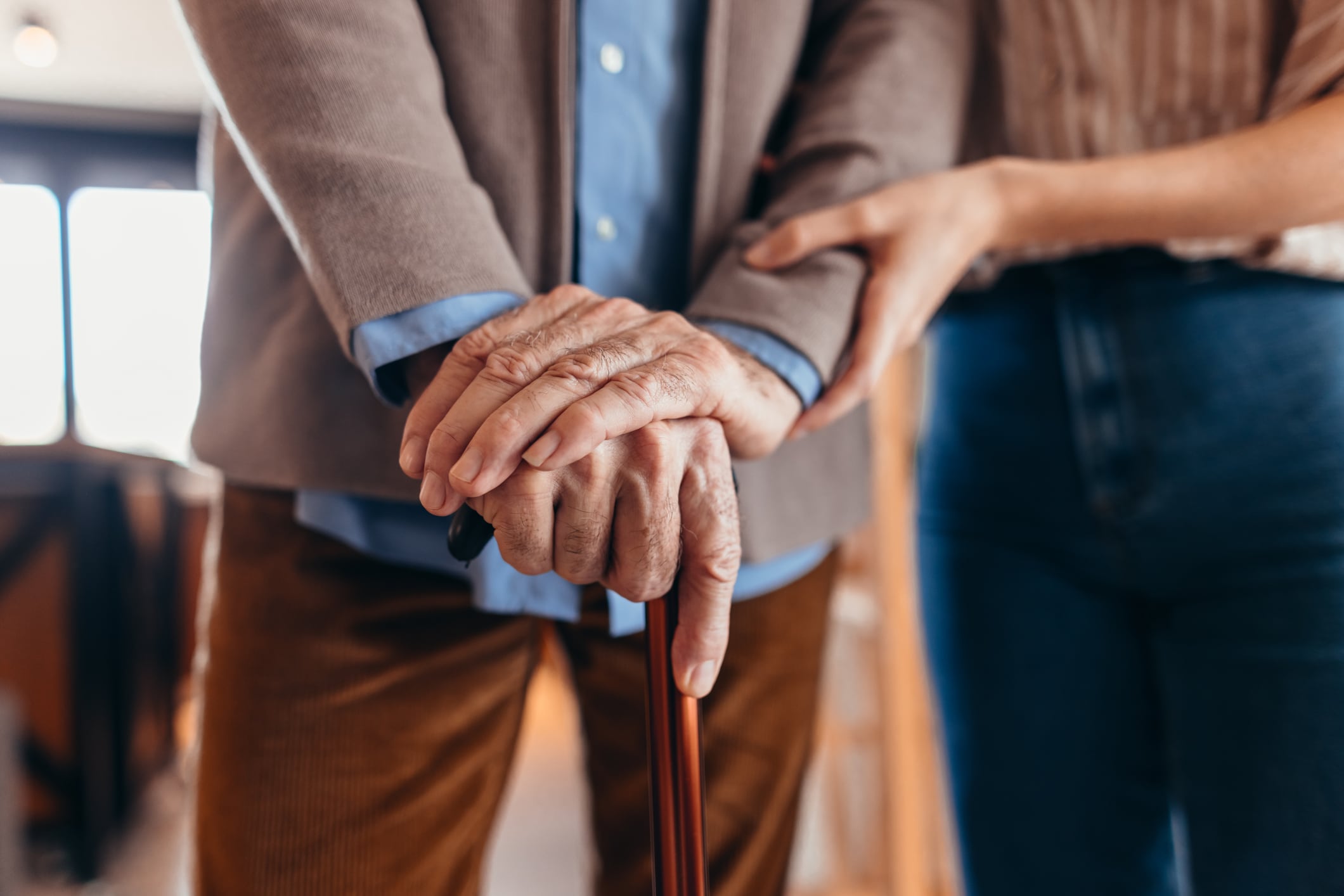 Close-up of a wrinkled hand holding a walking stick, being gently supported by a younger person, providing care and assistance to a senior adult