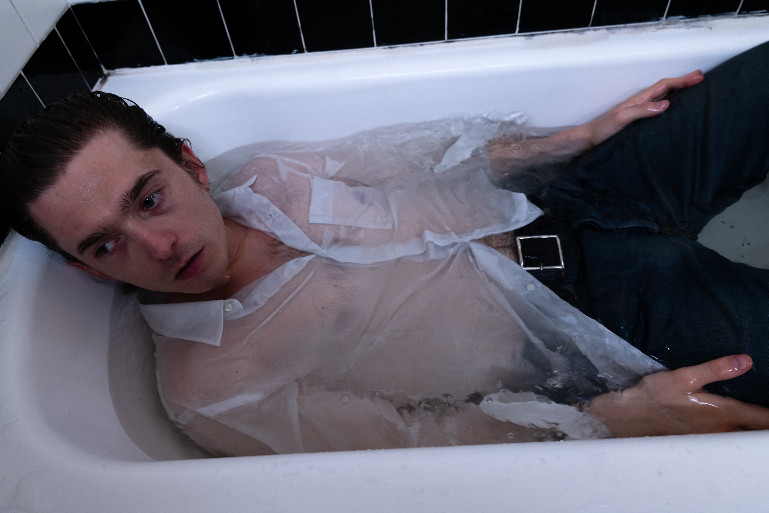 Austin Abrams lies down in a filled bathtub wearing a sheer button-down and dark jeans.