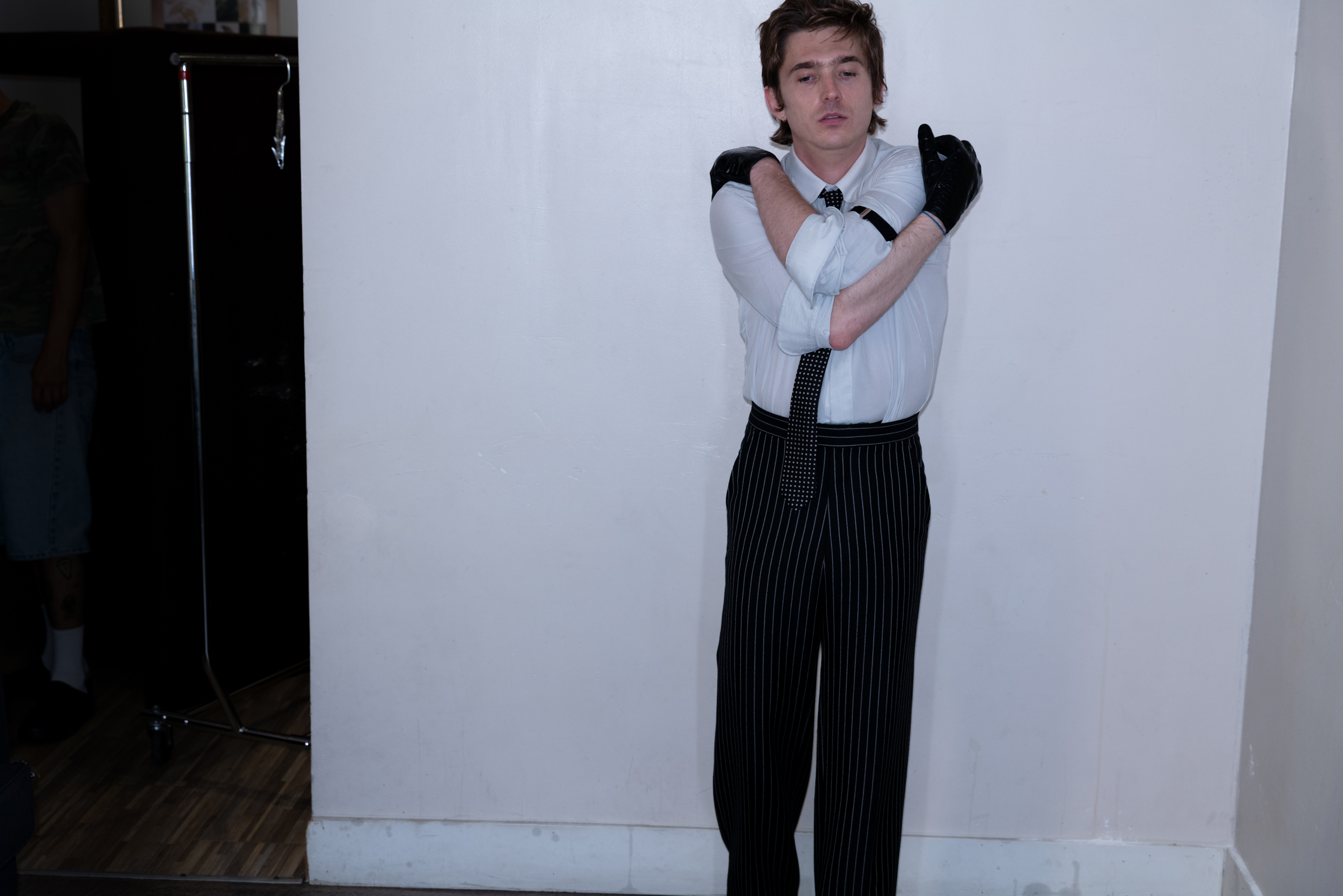Austin Abrams leans against a white wall, arms crossed over his chest.