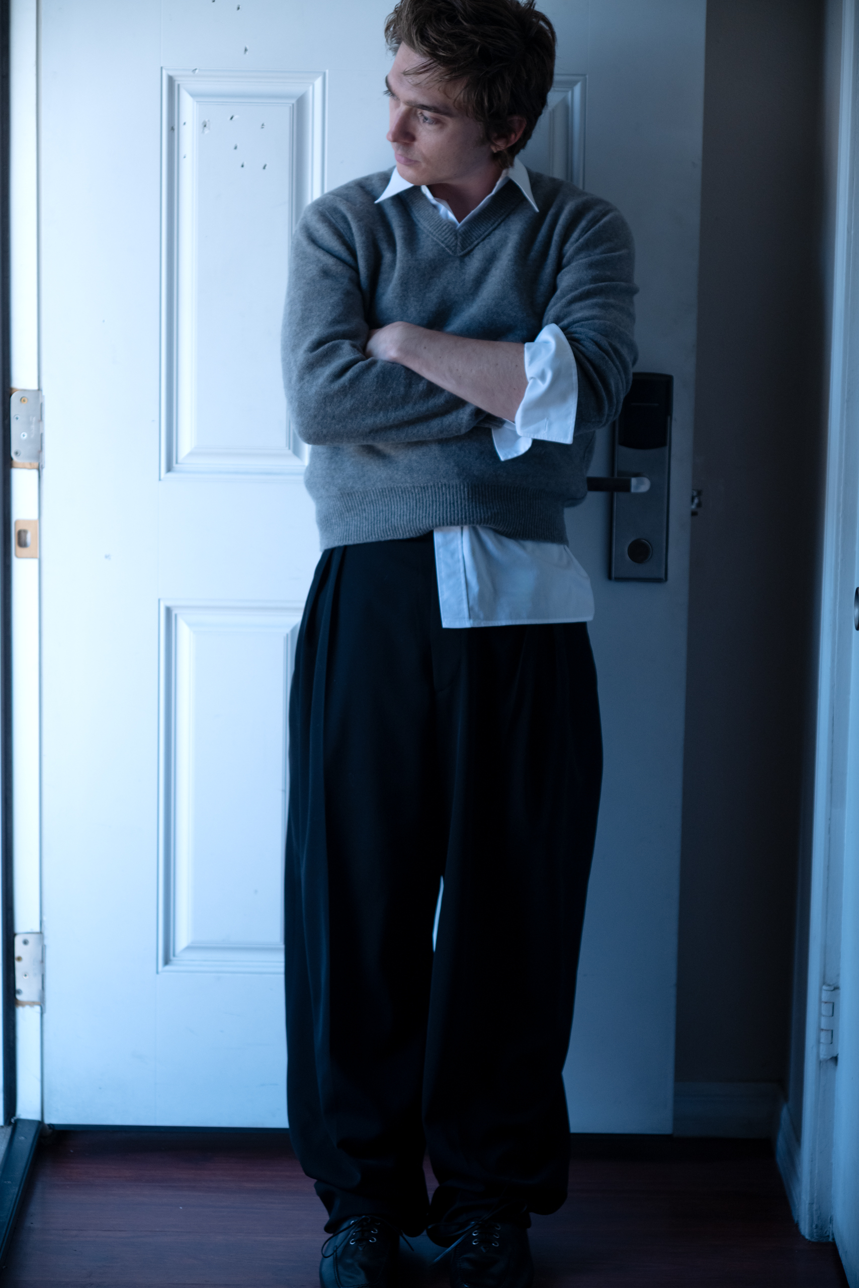 Austin Abrams leans against a white door, turning his head away from the camera, wearing a white button-down under a grey sweater with black slacks.