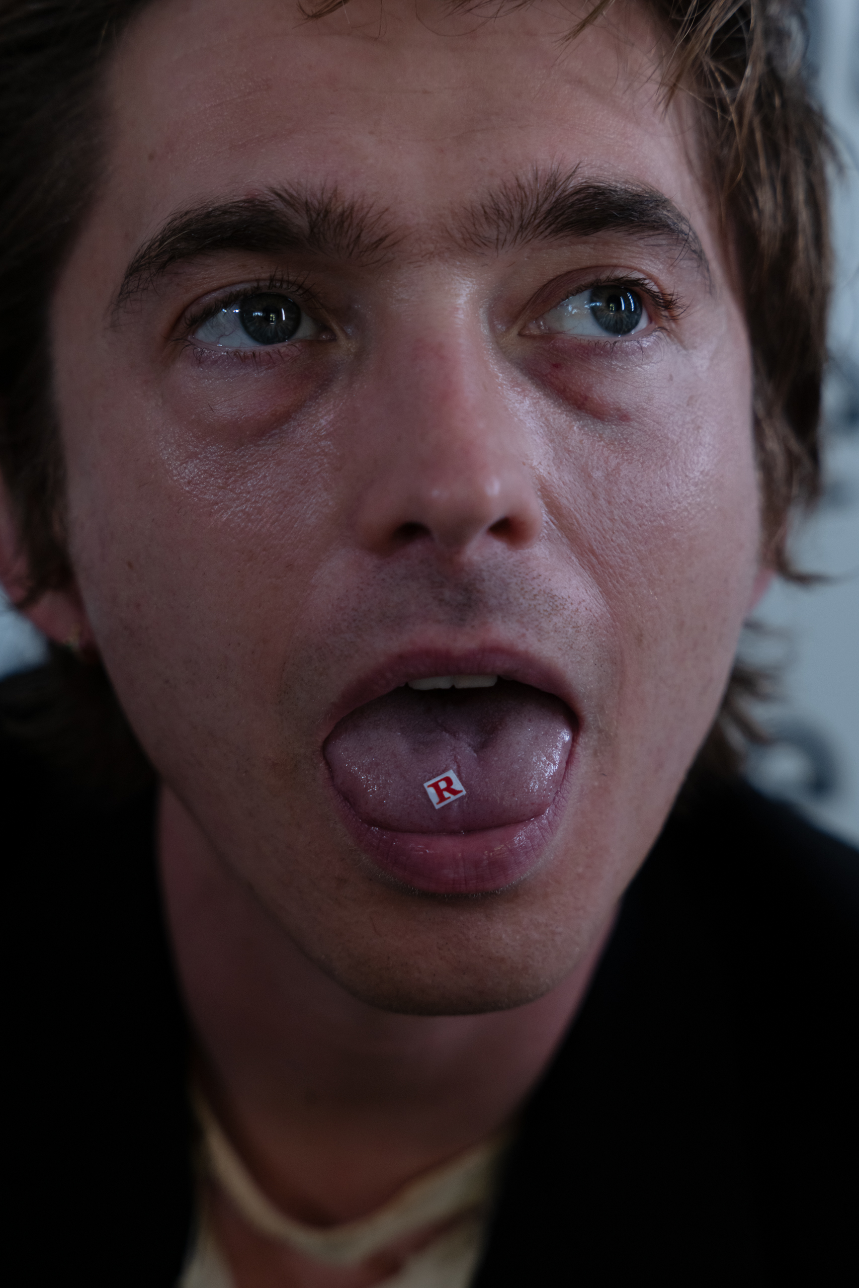 Austin Abrams poses with his tongue out, a small red letter "R" on it.