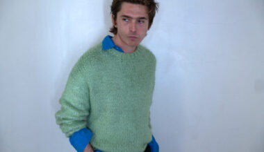 Austin Abrams turns his side to the camera, wearing a blue button-down under a green knit sweater.