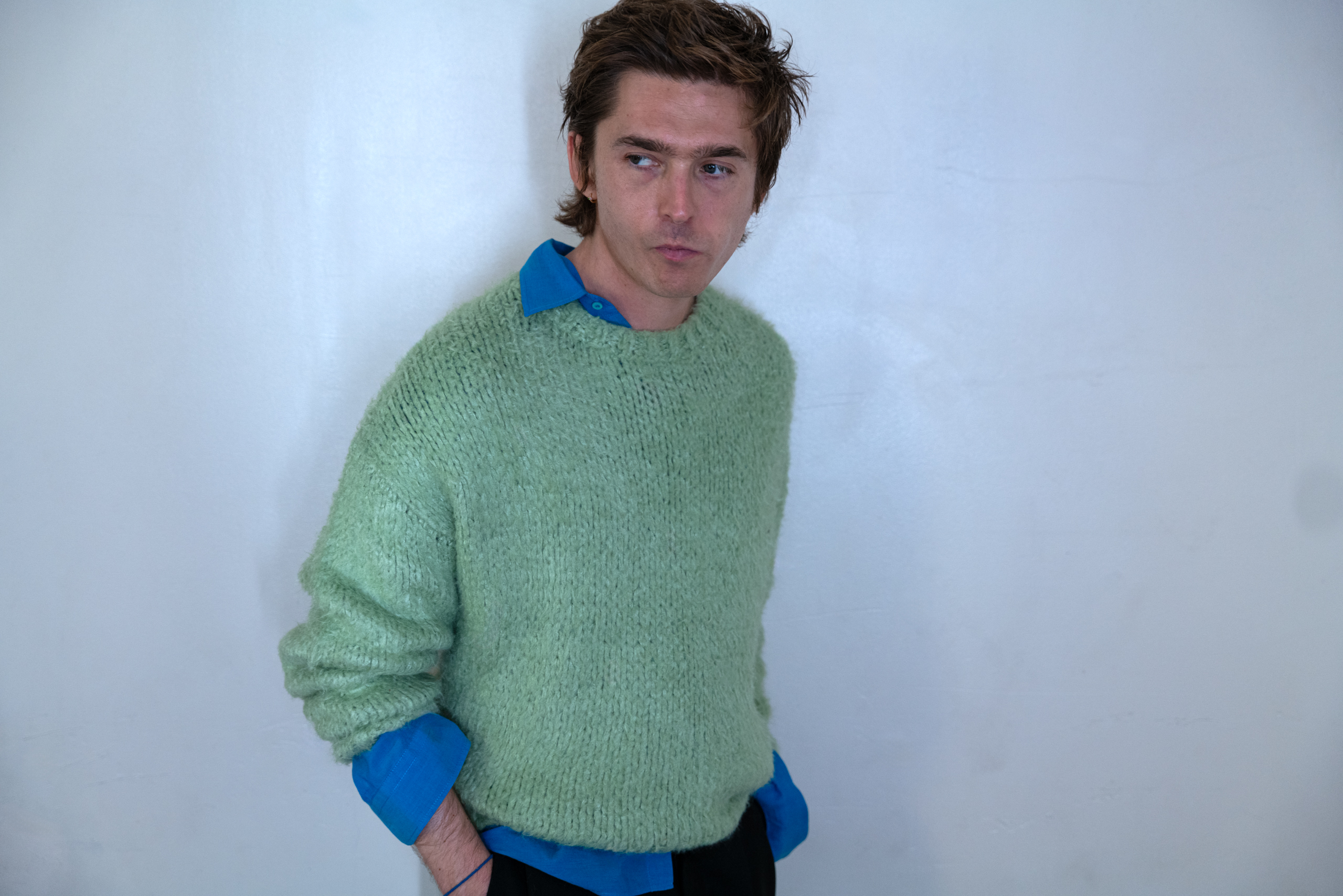 Austin Abrams turns his side to the camera, wearing a blue button-down under a green knit sweater.