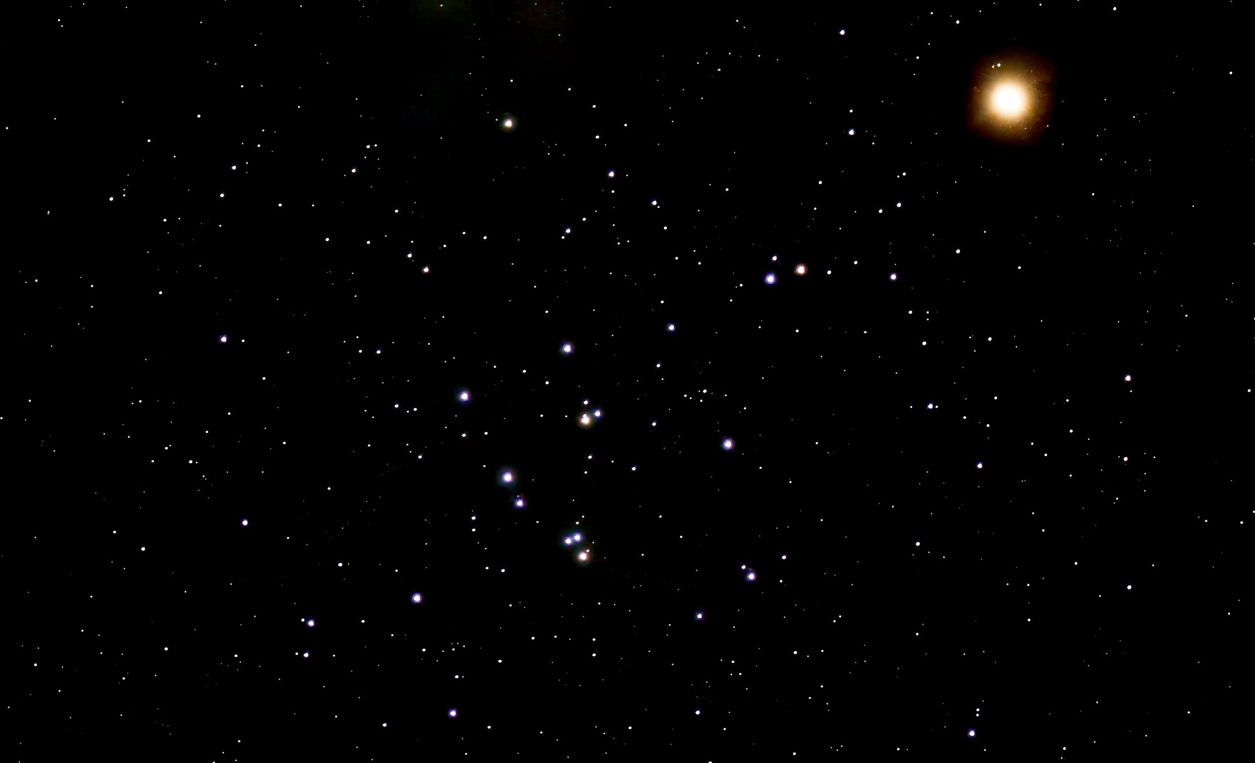 Photograph of the Beehive Cluster (M44) as a loose scattering of blue-white stars with a few orange stars across the lower center of the frame, set against a black sky filled with fainter stars. Mars appears at upper right as a bright, glowing orange-red disk.
