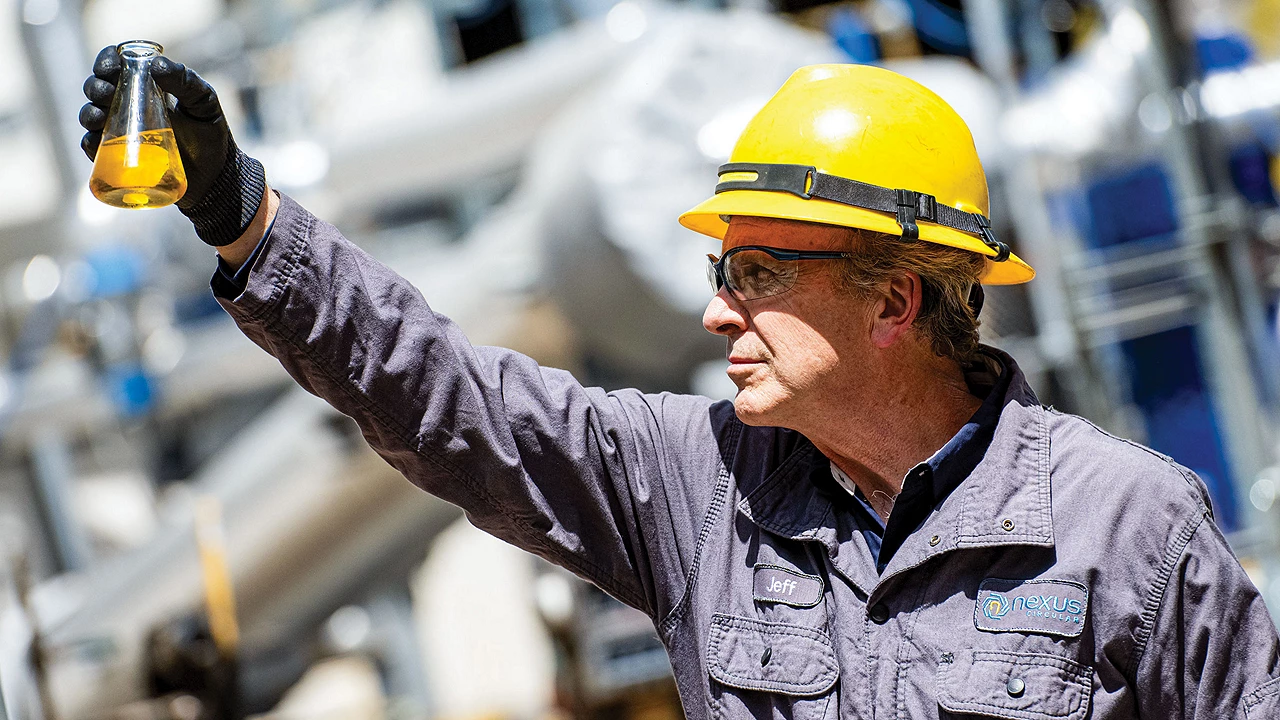 A man in a blue work suit examines a vial of liquid. His nametag on the left says “Jeff,” and the company name, Nexus Circular, is in blue lettering on the right.