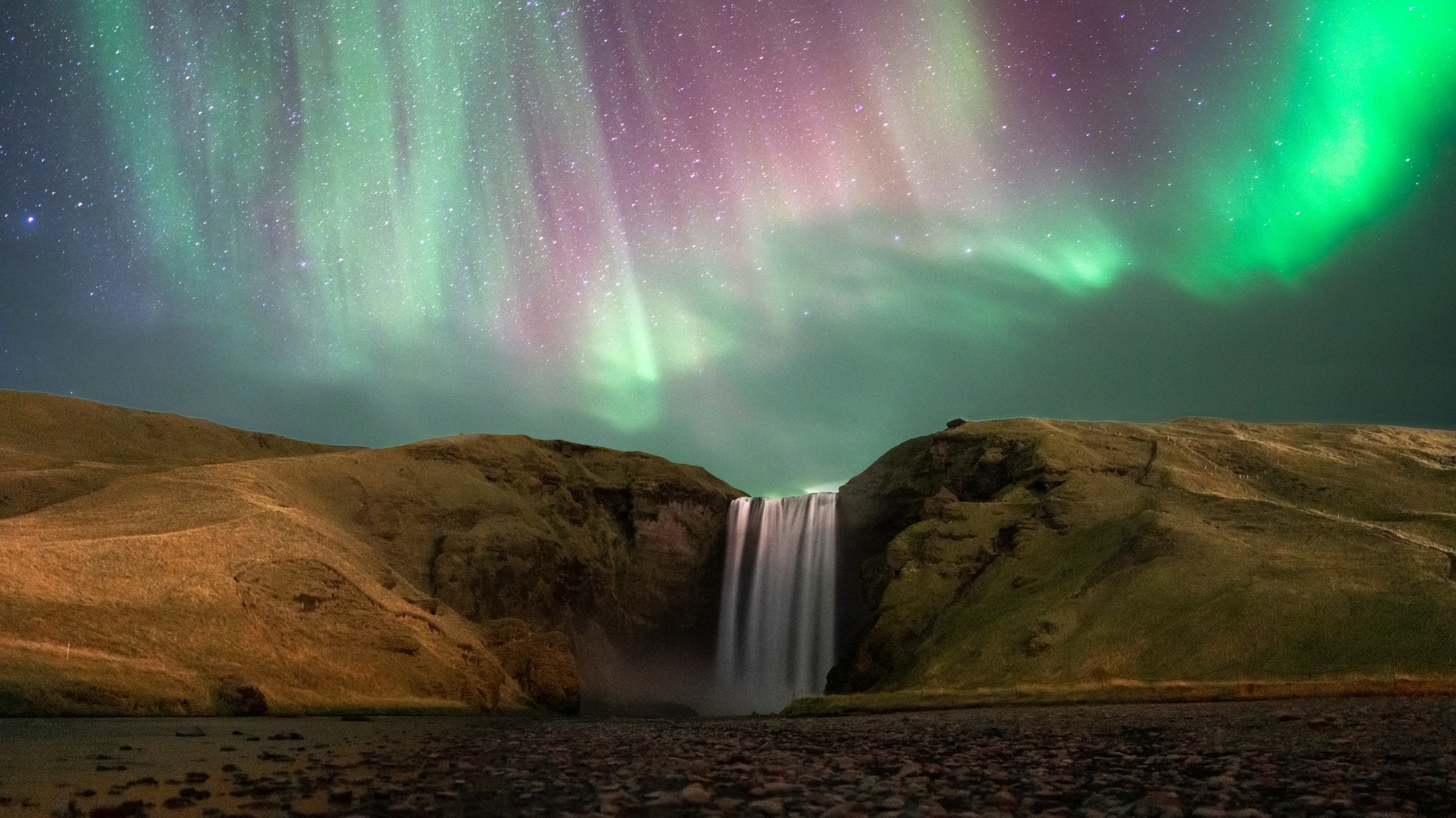 northern lights dance above a large waterfall in Iceland.