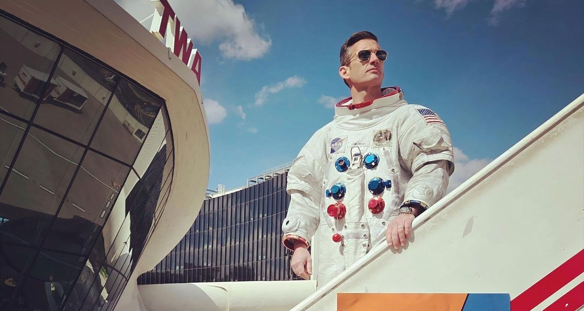 a man wearing a white spacesuit and sun glasses stands on a mobile stairway outside a 60's style airport terminal