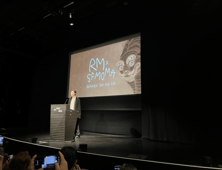 A person stands at a podium on stage in a dark auditorium, with a screen behind displaying “RM x SFMOMA: Between You and Me” and a drawing of a tiger. Audience members hold up phones.