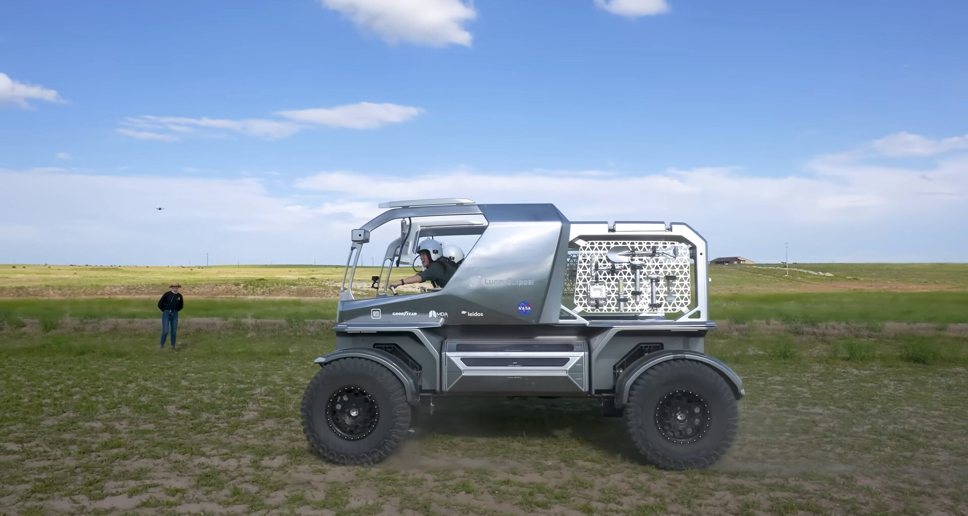 A shiny, silver vehicle with tools on a mesh rack on the back half and a large open cab up front drives across a grassy field under blue, partly cloudy skies. A person in a black shirt stands in the background with a drone hovering above their head. Two women in white helmets are piloting the vehicle from the cab.