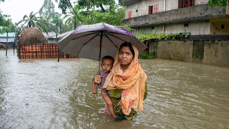 A woman carries her young child while wading through floodwaters week of heavy rain and mountain runoff in Bangladesh, July, 2026.